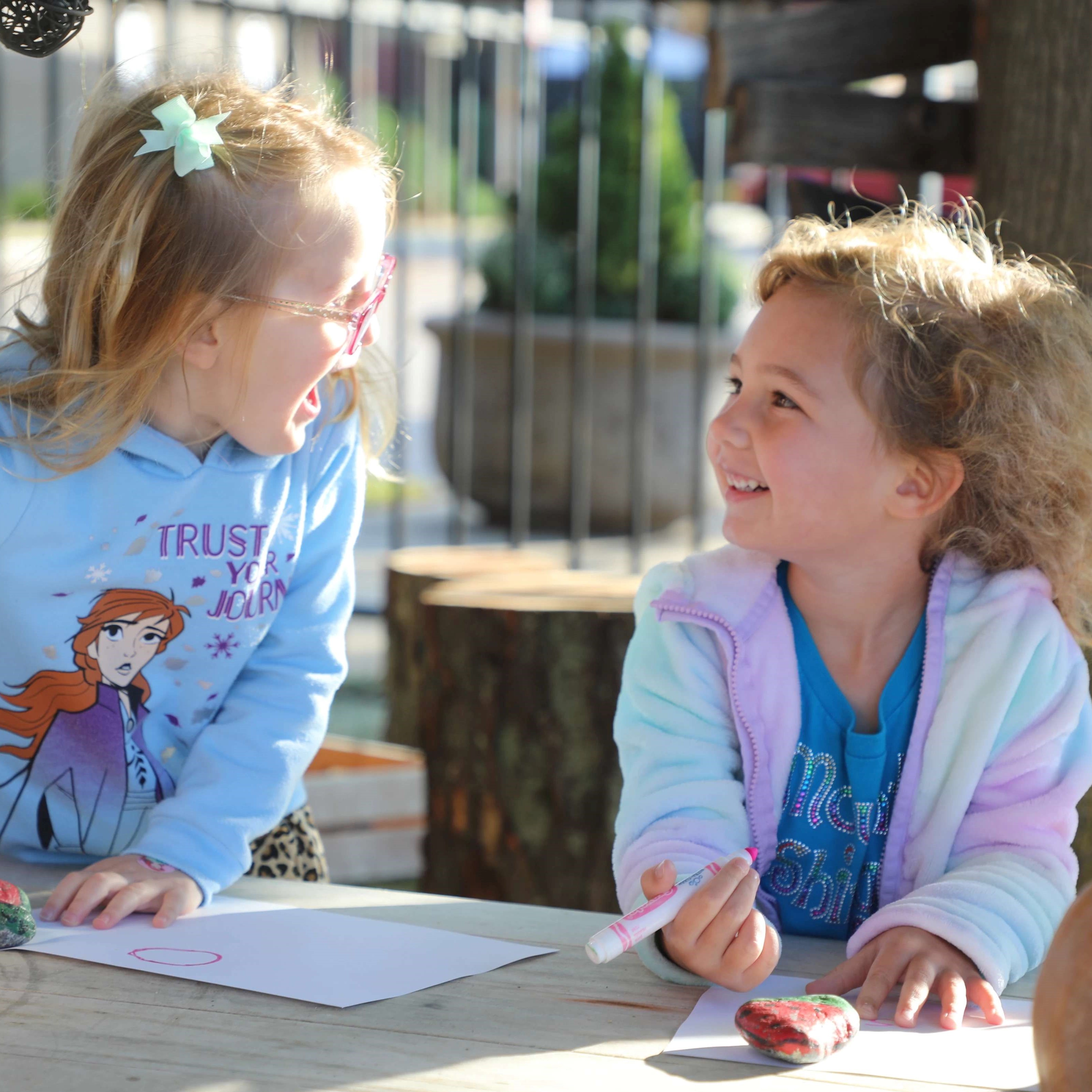 Two young girls smile and laugh at each other as they draw outside
