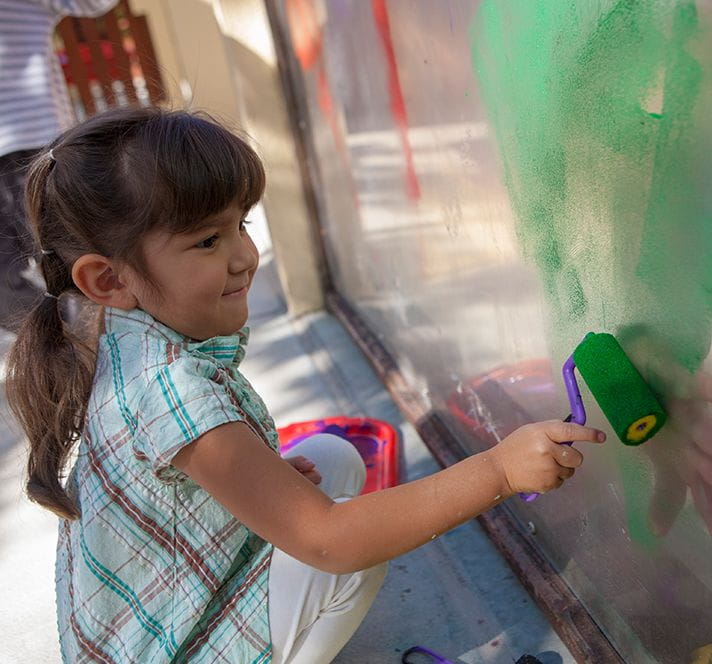 Kindergarten Prep girl with brown hair using a paint roller to paint a mural outside