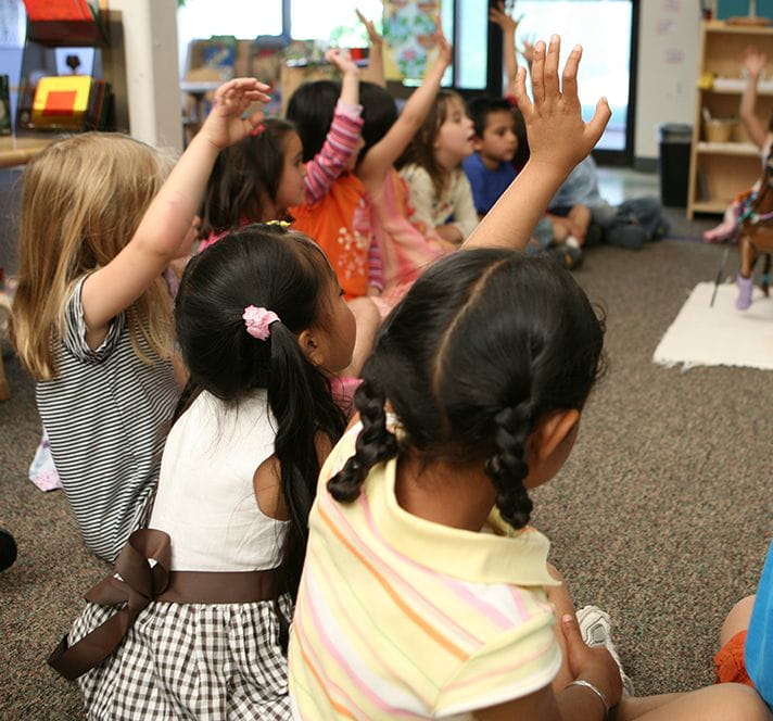 Group of Kindergarten Prep students sitting on the floor in a circle and raising their hands