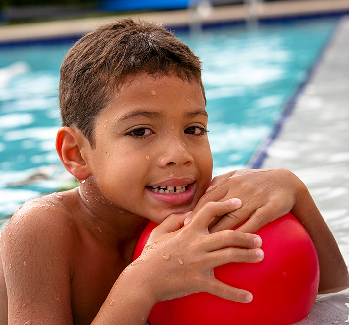 Kindergarten aged boy in a pool hanging on the edge with a red ball in hand