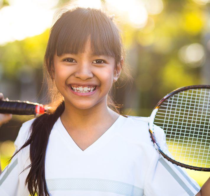 School aged girl with brown hair and bangs in a tennis uniform standing outside with a racquet