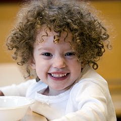 Preschool girl with short, brown, curly hair sitting at a table and smiling