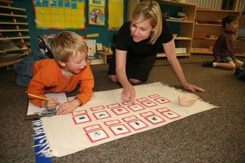 A teacher shows different cards on a mat to a young boy taking notes | Bright Horizons | Childcare