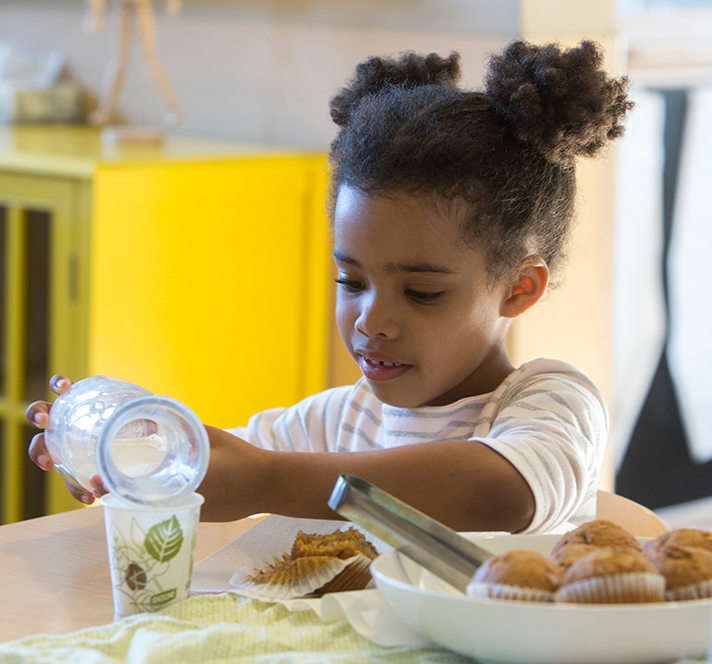 Preschool girl with two pigtails pouring milk into a cup next to a bowl of muffins